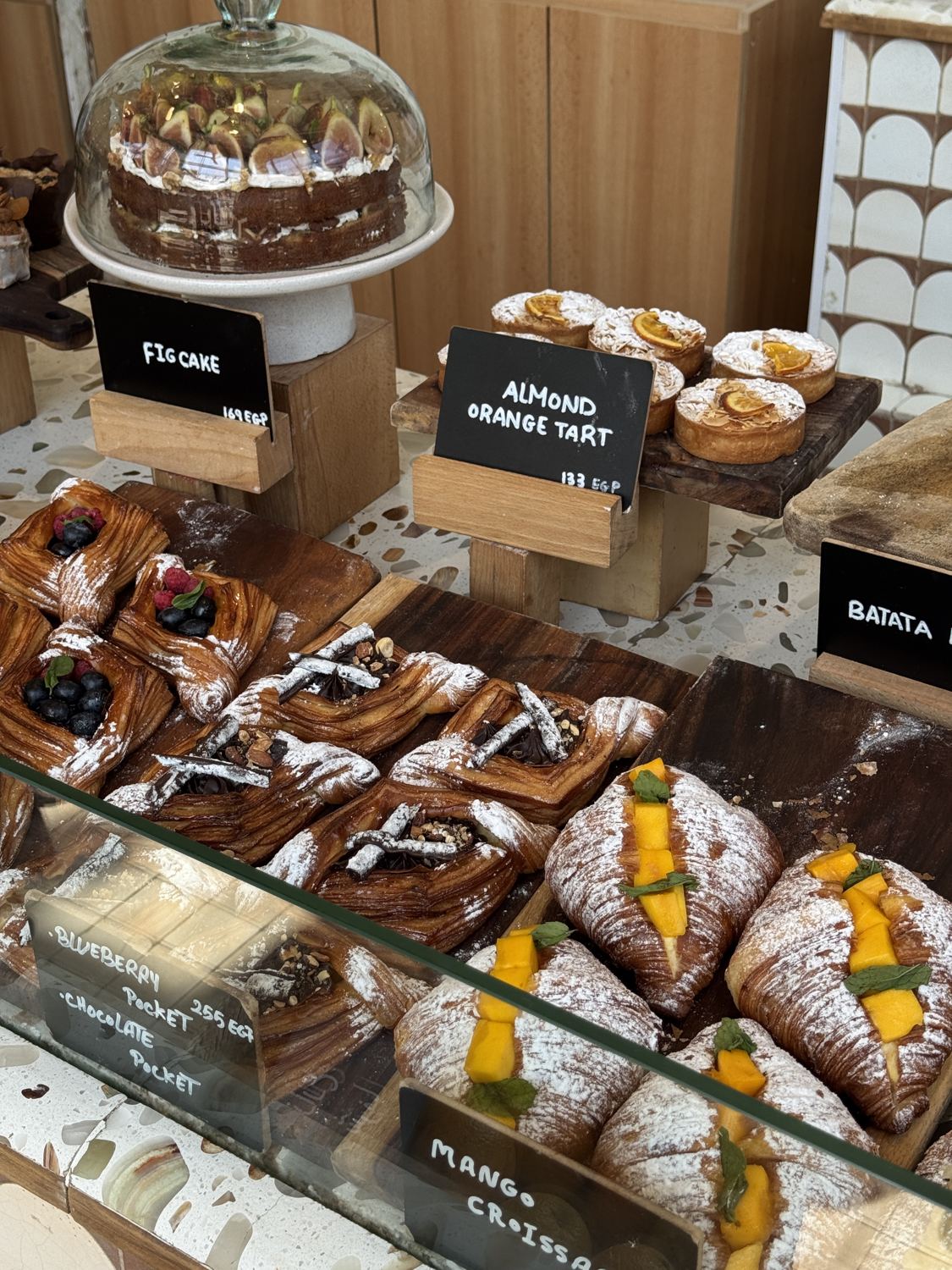 Bakery counter with fig cake under a glass dome, almond orange tarts and mango croissants