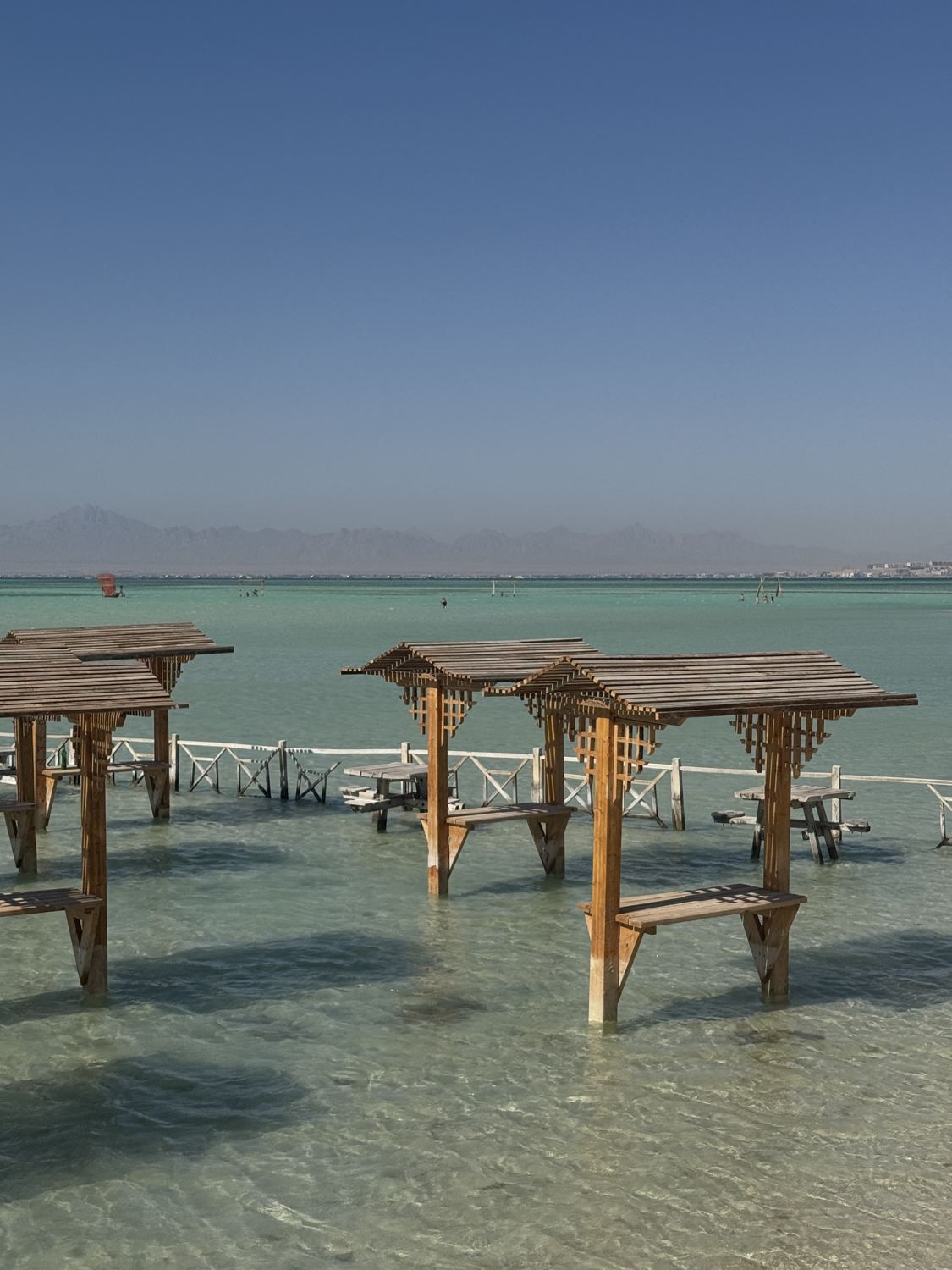 Wooden shade huts with benches standing in shallow turquoise water, with desert mountains across the sea