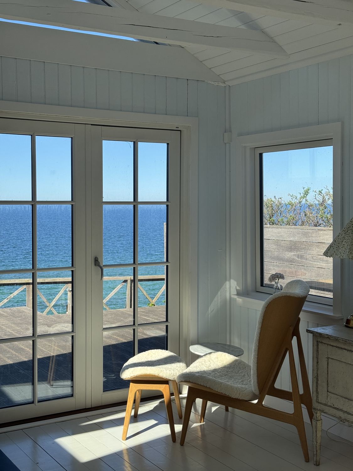 Wooden lounge chair with sheepskin by French doors overlooking a blue sea