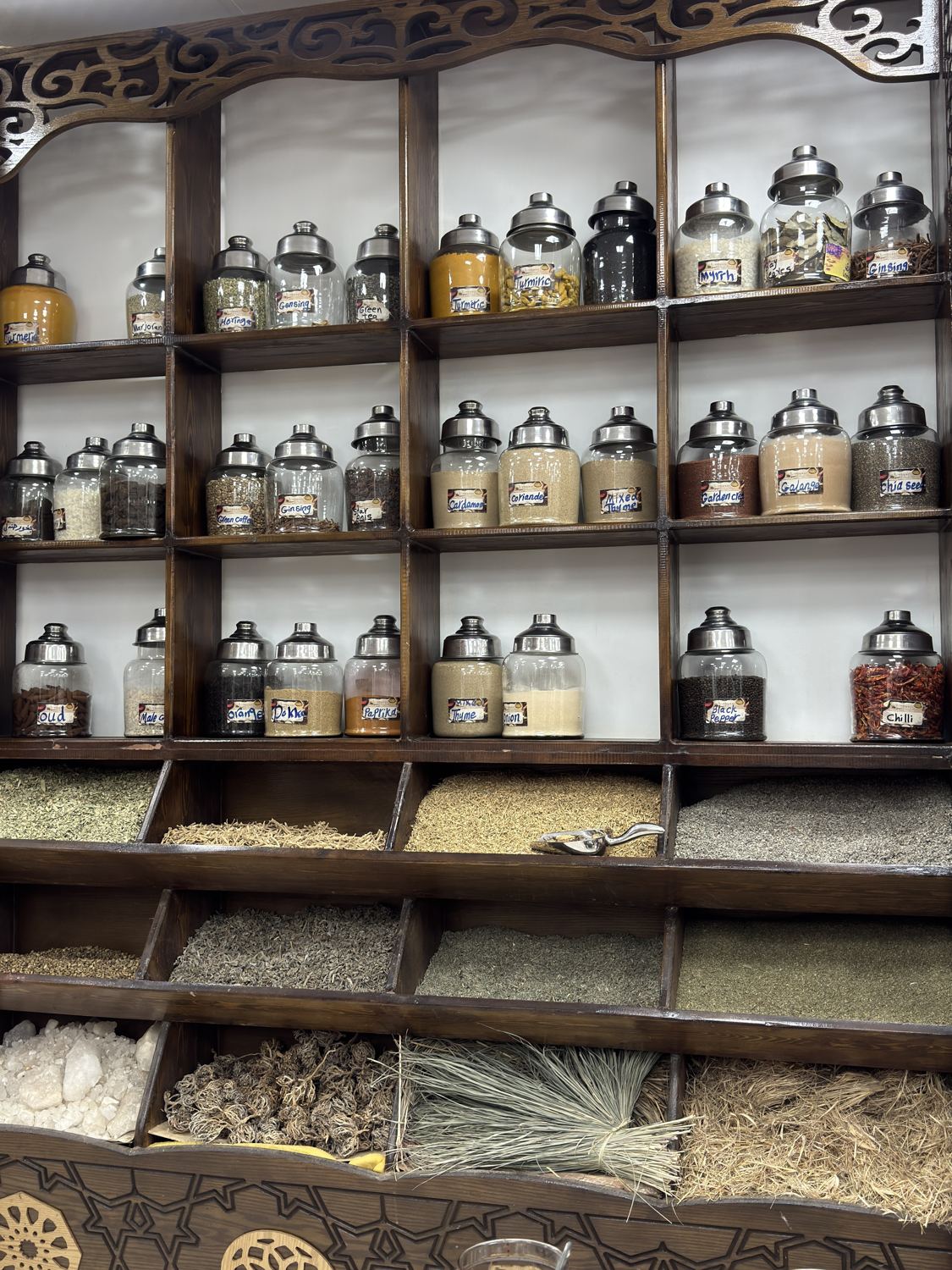 Wooden shelves of glass jars filled with spices and herbs, with dried herbs in bins below