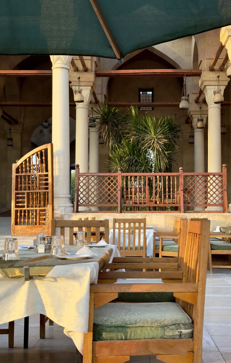 Wooden chairs and white tablecloths on a terrace in front of stone columns and mashrabiya screens